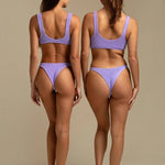 Two models wearing KABE SHOP INC Ginny bikini tops in textured lavender fabric, back view showing sporty silhouette.