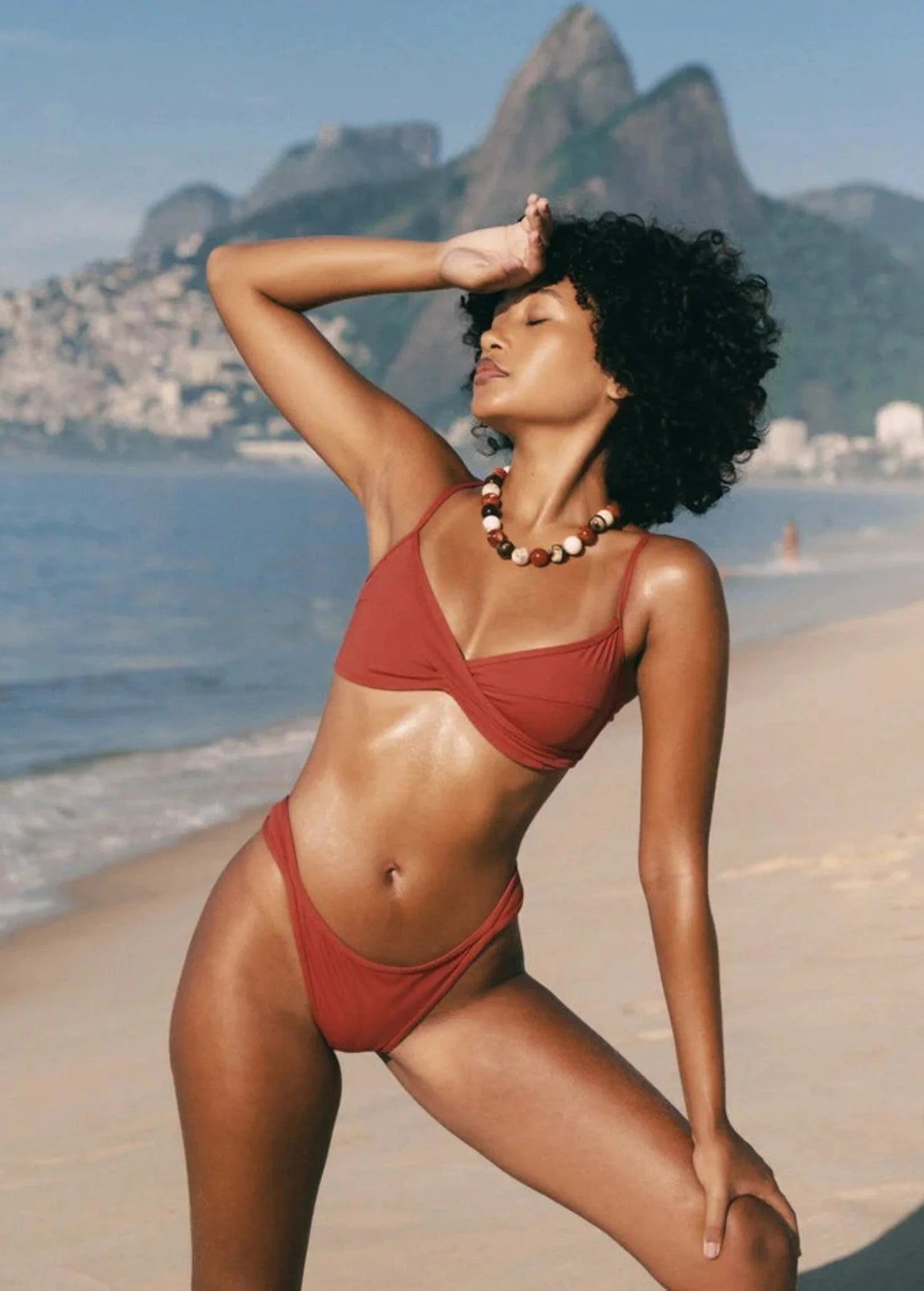 KABE SHOP INC Infinity Top in rust red with a front twist detail, worn by a model on a sunny beach.