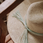 KABE SHOP INC Isabella wide brim straw hat with a decorative light green and white tassel detail on a wooden chair.