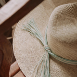 KABE SHOP INC Isabella wide brim straw hat with a decorative light green and white tassel detail on a wooden chair.