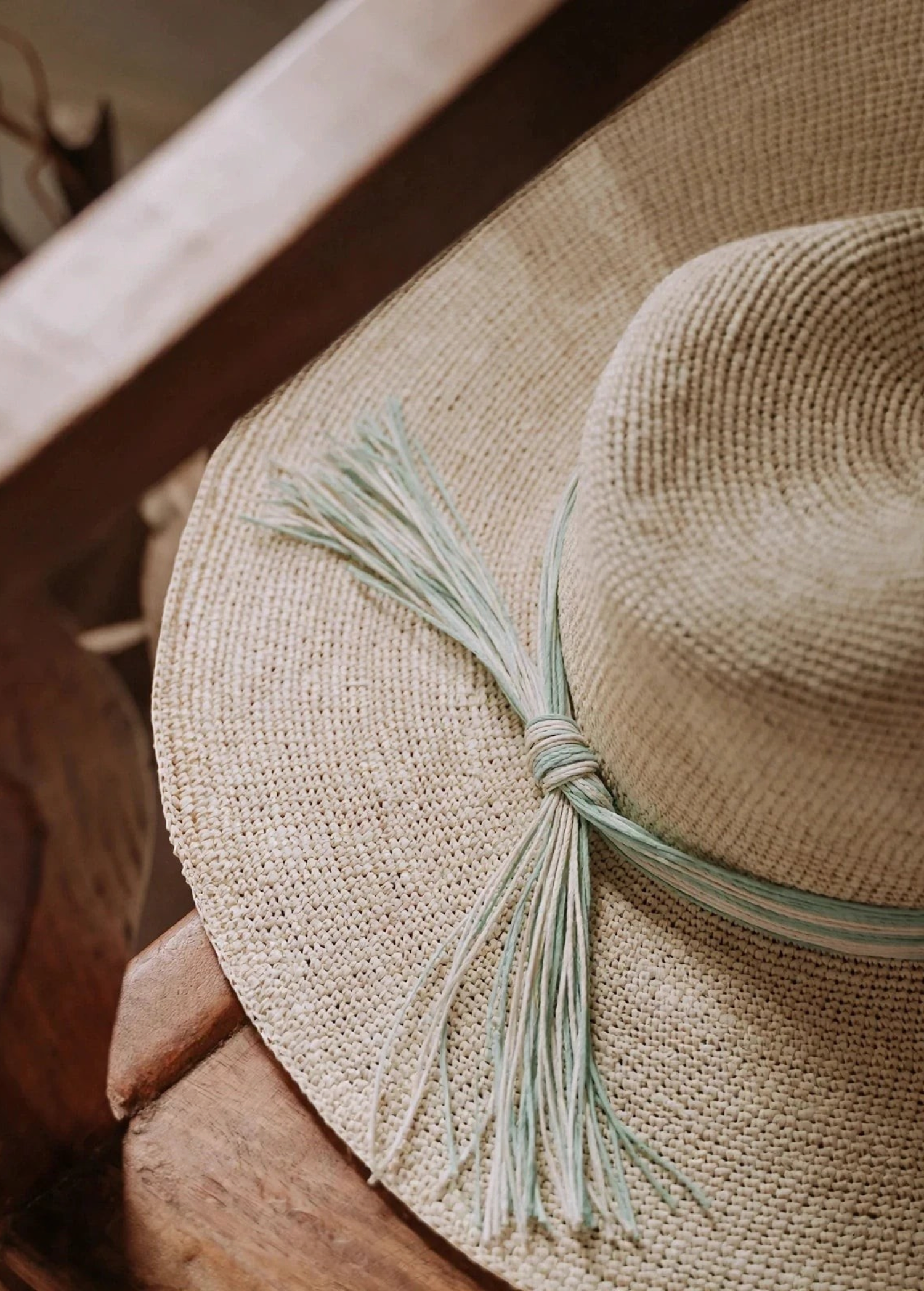KABE SHOP INC Isabella wide brim straw hat with a decorative light green and white tassel detail on a wooden chair.