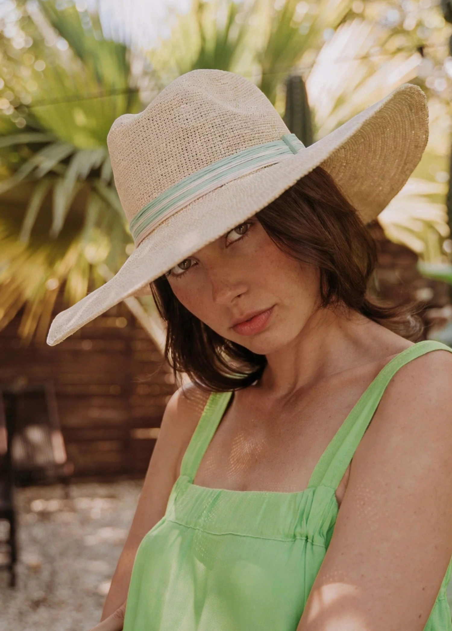 KABE SHOP INC Isabella wide brim straw hat with a light green striped ribbon, worn by a woman in a sunny garden.