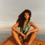 KABE SHOP INC Magdalena Pareo with a fluid landscape print worn by a woman sitting on a rusted structure by the sea.