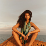 KABE SHOP INC Magdalena Pareo with a fluid landscape print worn by a woman sitting on a rusted structure by the sea.