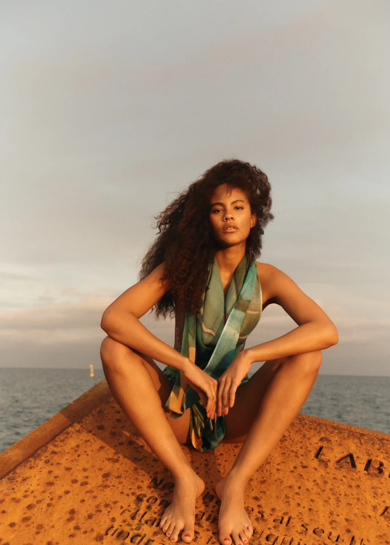 KABE SHOP INC Magdalena Pareo with a fluid landscape print worn by a woman sitting on a rusted structure by the sea.