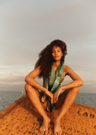 KABE SHOP INC Magdalena Pareo with a fluid landscape print worn by a woman sitting on a rusted structure by the sea.