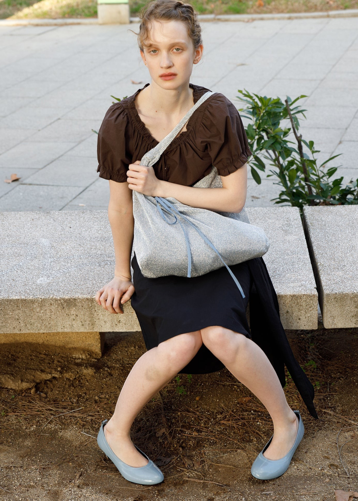 Model sitting on a stone bench wearing a KABE SHOP INC Deiji Studios metallic knit chain bag with a drawstring pouch.