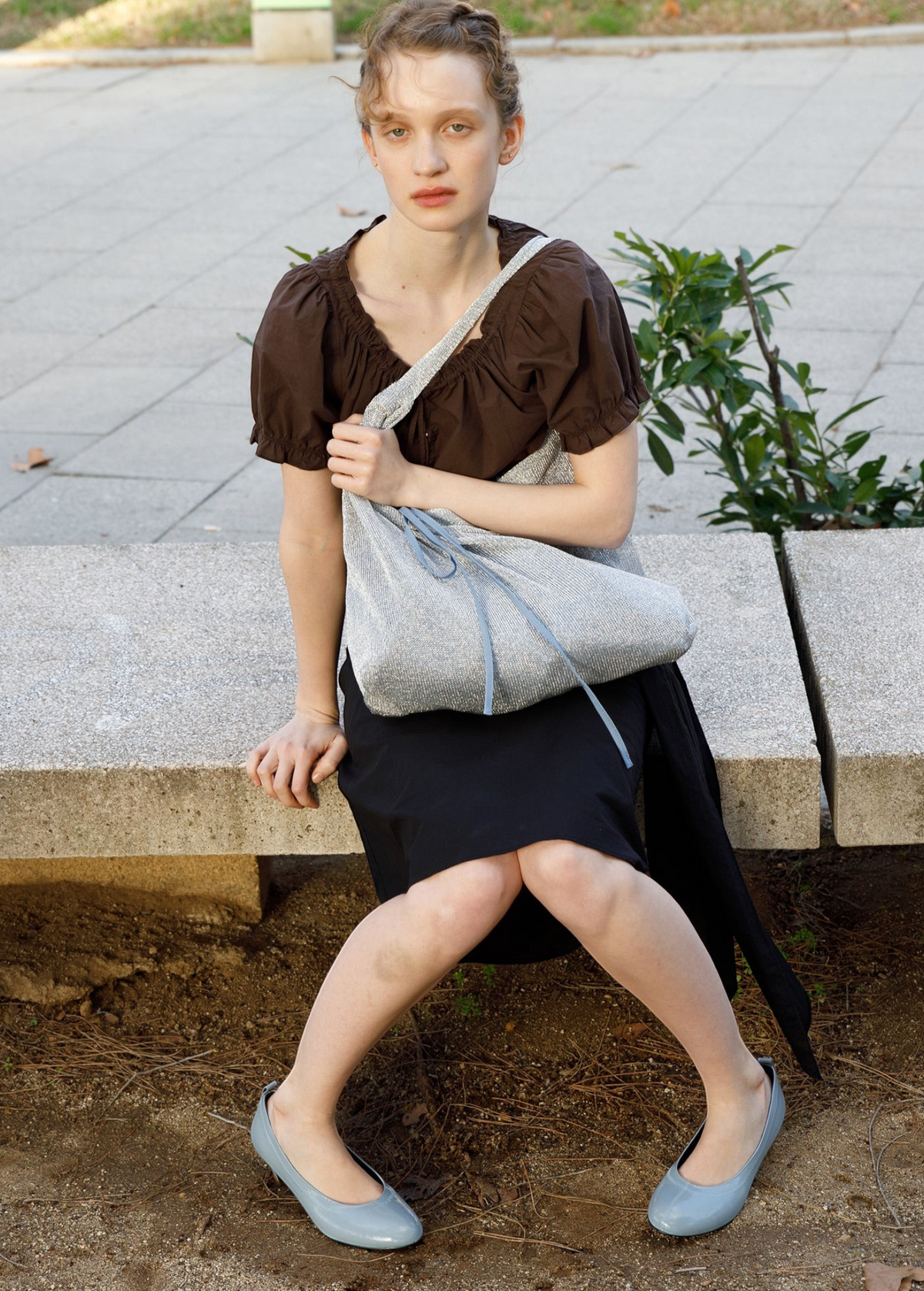 Model sitting on a stone bench wearing a KABE SHOP INC Deiji Studios metallic knit chain bag with a drawstring pouch.