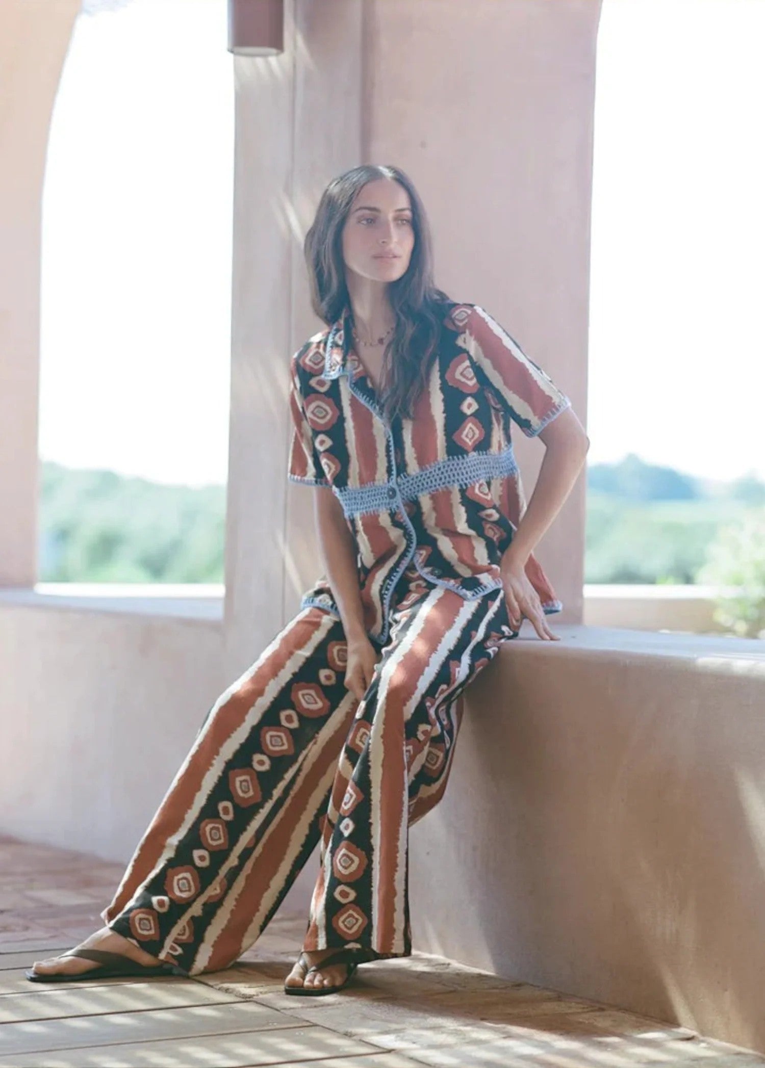 Model wearing KABE SHOP INC Nera wide leg pants with a tribal ikat print in brown and cream on a sunny terrace.