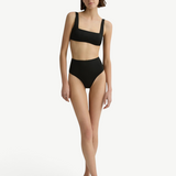 Model wearing KABE SHOP INC Poppy high-waisted black bikini bottoms and matching crop top against a white wall.