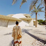 KABE SHOP INC The Sharis Kimono in beige and brown tie-dye worn by a woman with a bun, standing near modern architecture