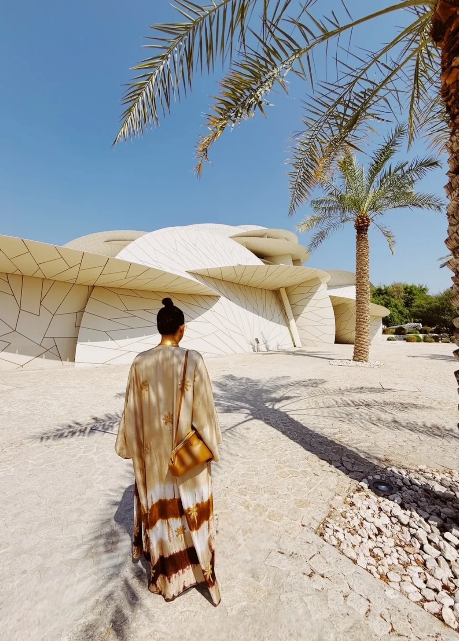 KABE SHOP INC The Sharis Kimono in beige and brown tie-dye worn by a woman with a bun, standing near modern architecture