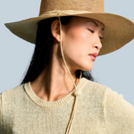 Model wearing a KABE SHOP INC Vitoria wide brim straw hat with a chin strap against a blue background