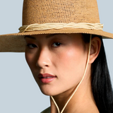 A model wearing the KABE SHOP INC Vitoria wide brim straw hat with a chin strap against a blue background.