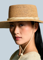 A model wearing the KABE SHOP INC Vitoria wide brim straw hat with a chin strap against a blue background.