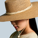 KABE SHOP INC Vitoria wide brim straw hat in tan with a cream cord chin strap worn by a woman against a blue backdrop