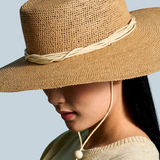 KABE SHOP INC Vitoria wide brim straw hat in tan with a cream cord chin strap worn by a woman against a blue backdrop