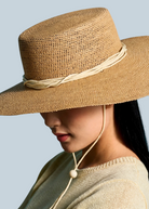 KABE SHOP INC Vitoria wide brim straw hat in tan with a cream cord chin strap worn by a woman against a blue backdrop