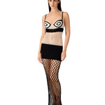 A woman stands against a plain white background wearing the La Femme Dress—featuring a black-and-white crochet fabric with circular patterns, styled with a black mini skirt lining and a sheer fishnet maxi skirt layered over sandals.