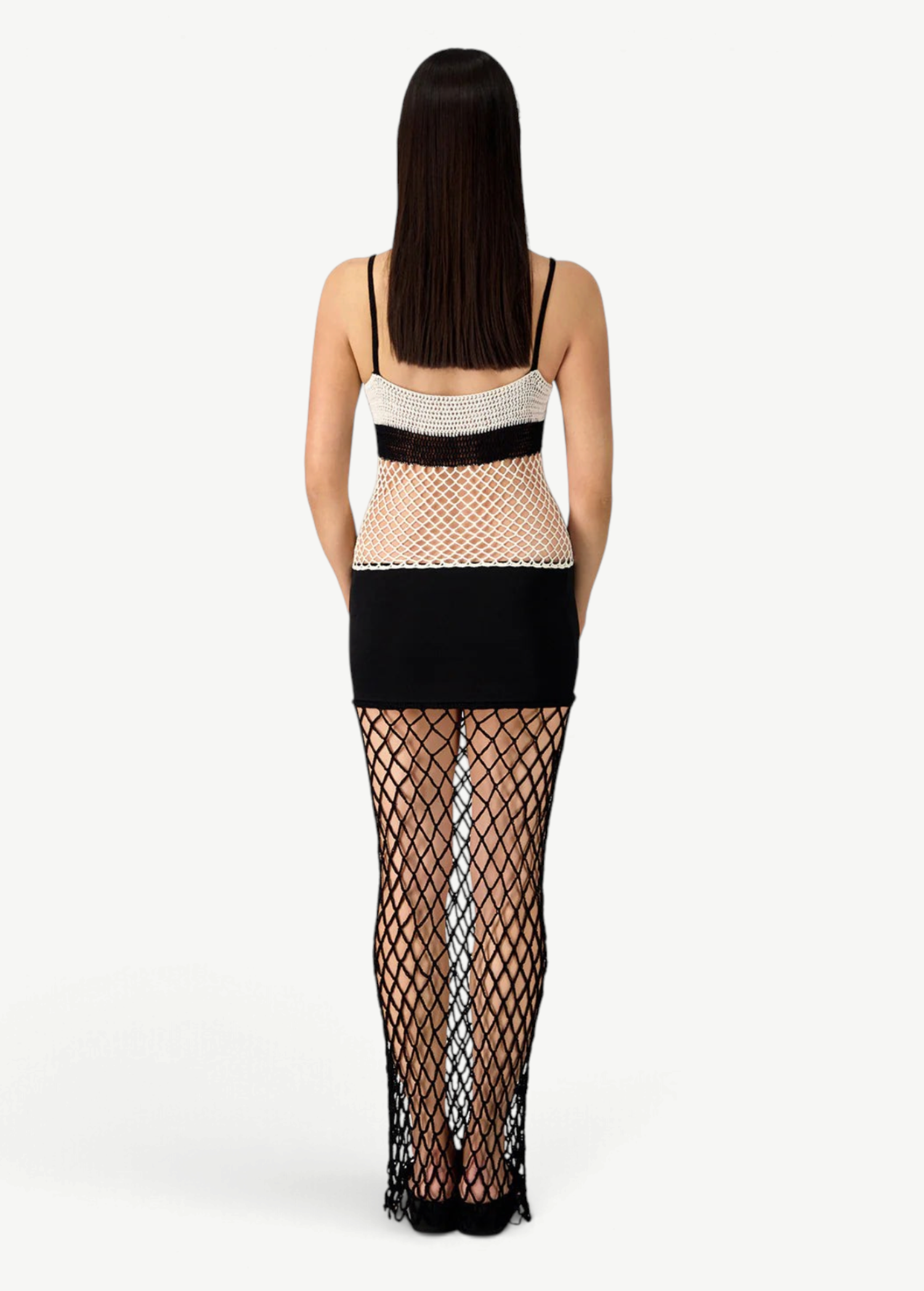 A woman with long brown hair stands facing away, wearing the La Femme Dress—a sleeveless style featuring a beige and black mesh top, a black mini skirt, and a long black fishnet overlay with sheer textures covering her legs. Set against a white background.