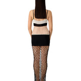 A woman with long brown hair stands facing away, wearing the La Femme Dress—a sleeveless style featuring a beige and black mesh top, a black mini skirt, and a long black fishnet overlay with sheer textures covering her legs. Set against a white background.
