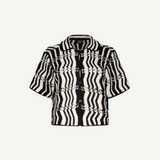 The Riviera Shirt is a black-and-white short-sleeve button-up summer shirt featuring a bold zigzag wavy pattern and a collar, displayed on a plain white background. This black-and-white crochet Riviera Shirt adds a unique textured twist to classic warm-weather style.