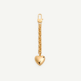 The Heart Bag Charm features a shiny, puffy heart pendant hanging from a 14KT gold plated chain, adding timeless elegance to any accessory. Shown on a plain white background, this piece channels the chic style of a Martha Calvo pendant.