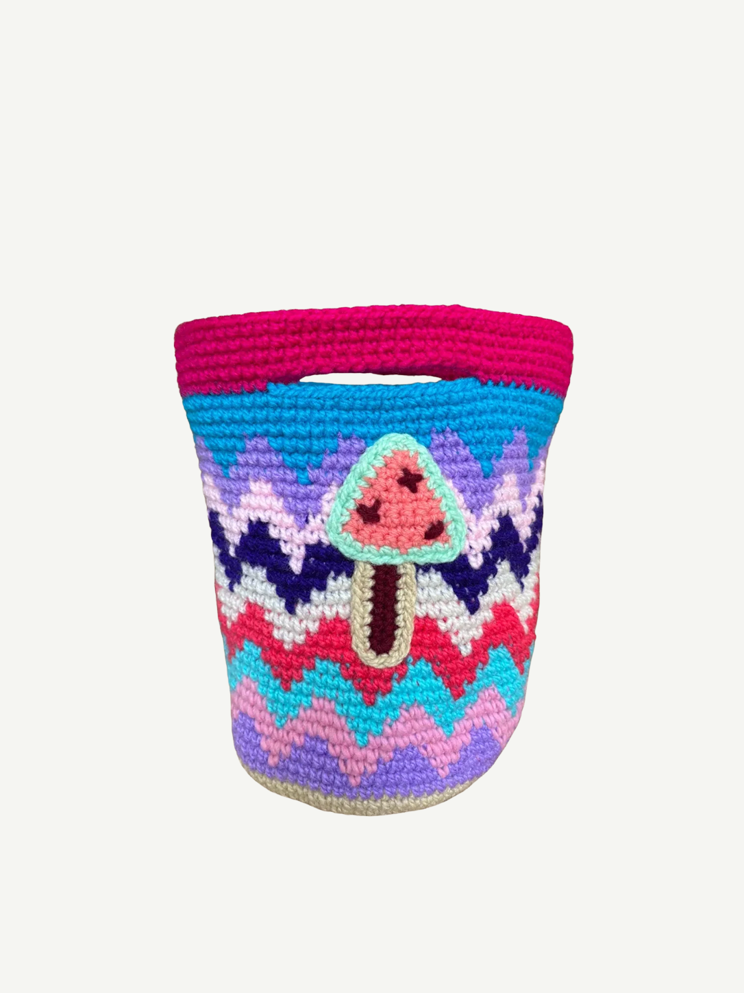 Introducing the Champinones Bag: a vibrant, handmade crocheted basket with a zigzag pattern in pink, blue, purple, and white. Expertly crafted from crochet wool, it features an adorable applique of a pink mushroom with a brown stem on the front. This unique piece infuses any space with playful charm and artistic flair.