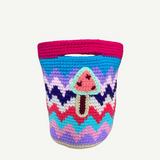 Introducing the Champinones Bag: a vibrant, handmade crocheted basket with a zigzag pattern in pink, blue, purple, and white. Expertly crafted from crochet wool, it features an adorable applique of a pink mushroom with a brown stem on the front. This unique piece infuses any space with playful charm and artistic flair.