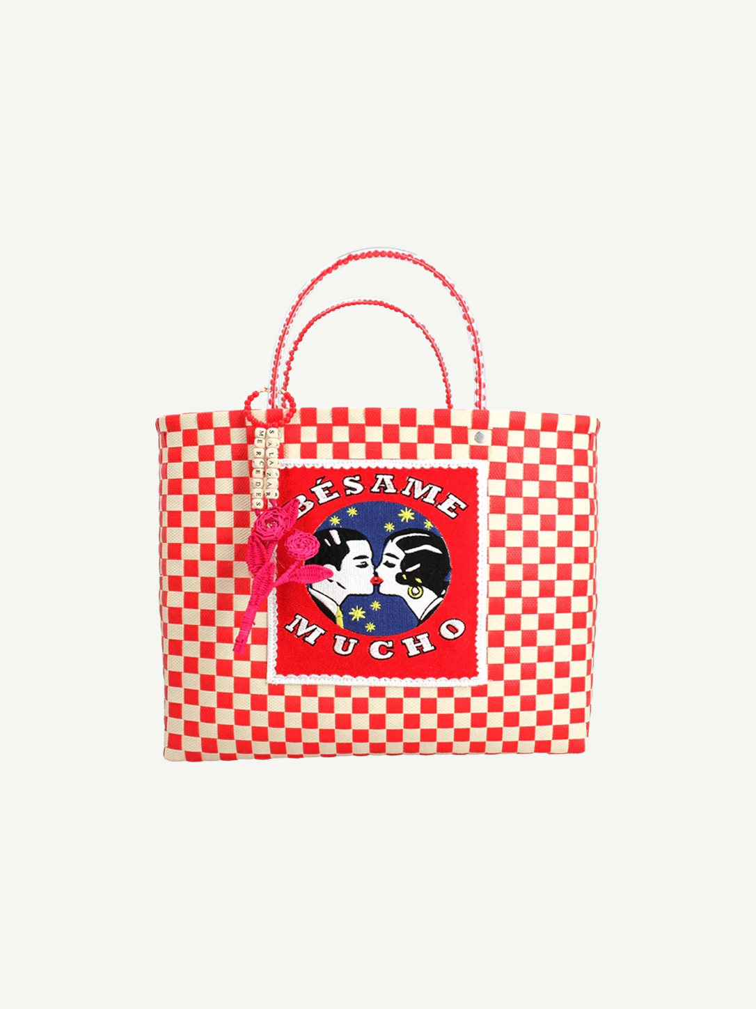 Introducing the Besame Mucho Medium Handbag: a charming handcrafted accessory featuring red and white checkered fabric, beaded handles, a pink bow accent, and a unique patch displaying two faces with the words “Bésame Mucho.” This playful handbag adds an irresistible touch of whimsy to any outfit.