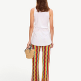 A person from the back wearing a white sleeveless top and Ophelia Knit Pant flared crochet pants with a colorful vertical chevron pattern, holding a small straw handbag.