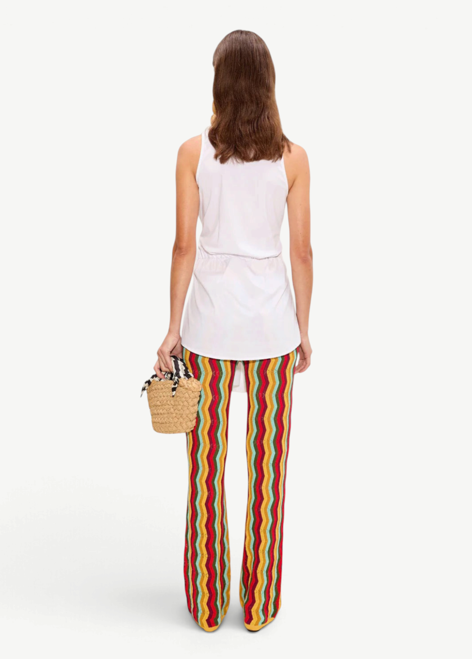 A person from the back wearing a white sleeveless top and Ophelia Knit Pant flared crochet pants with a colorful vertical chevron pattern, holding a small straw handbag.