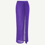 The Sarong Skirt is a long, sheer, pleated purple skirt featuring a side slit and a draped tie detail at the waist, shown against a plain white background.