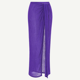 The Sarong Skirt is a long, sheer, pleated purple skirt featuring a side slit and a draped tie detail at the waist, shown against a plain white background.