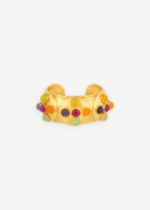 Introducing the Theodora Cuff: a wide gold ring with a gently pointed front, this handcrafted piece is adorned with small, colorful round gemstones—including red, green, purple, and orange—set against a simple white backdrop. Effortlessly wearable, the Theodora Cuff brings vibrant elegance to any ensemble.