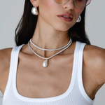 Woman wearing pink-tinted heart-shaped sunglasses, the Jordyn Hoops in Rhodium with a pierced back, layered silver necklaces with a pendant, and a white ribbed tank top, posing against a plain light background.