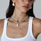 Woman wearing pink-tinted heart-shaped sunglasses, the Jordyn Hoops in Rhodium with a pierced back, layered silver necklaces with a pendant, and a white ribbed tank top, posing against a plain light background.