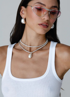 Woman wearing pink-tinted heart-shaped sunglasses, the Jordyn Hoops in Rhodium with a pierced back, layered silver necklaces with a pendant, and a white ribbed tank top, posing against a plain light background.