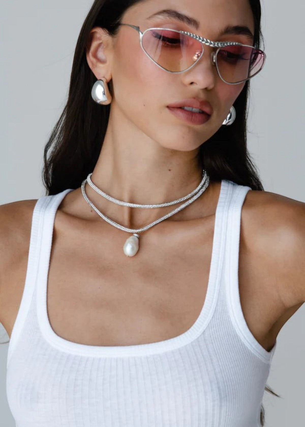 Woman wearing pink-tinted heart-shaped sunglasses, the Jordyn Hoops in Rhodium with a pierced back, layered silver necklaces with a pendant, and a white ribbed tank top, posing against a plain light background.
