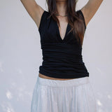 A woman wearing a white hat, the Sunnery Top in black Tencel jersey with a V-neck, and a white skirt stands with her arms raised behind her head against a plain light background.
