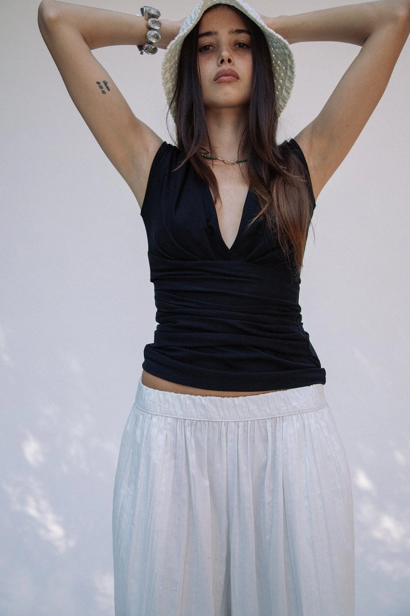 A woman wearing a white hat, the Sunnery Top in black Tencel jersey with a V-neck, and a white skirt stands with her arms raised behind her head against a plain light background.