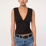 A person stands against a plain background, wearing the Sunnery Top—a sleeveless black V-neck crafted from soft Tencel jersey—paired with blue jeans, a black belt, and gold hoop earrings, looking straight at the camera with a neutral expression.