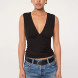 A person stands against a plain background, wearing the Sunnery Top—a sleeveless black V-neck crafted from soft Tencel jersey—paired with blue jeans, a black belt, and gold hoop earrings, looking straight at the camera with a neutral expression.