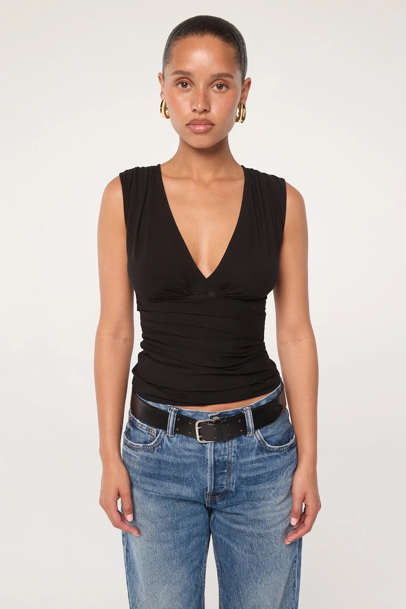 A person stands against a plain background, wearing the Sunnery Top—a sleeveless black V-neck crafted from soft Tencel jersey—paired with blue jeans, a black belt, and gold hoop earrings, looking straight at the camera with a neutral expression.