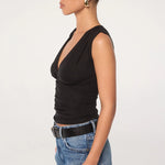 A woman in profile wears the Sunnery Top—a black v-neck made from soft Tencel jersey—paired with blue jeans, a black belt, and gold hoop earrings. Her hair is slicked back in a low bun, set against a plain white background.