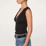 A woman in profile wears the Sunnery Top—a black v-neck made from soft Tencel jersey—paired with blue jeans, a black belt, and gold hoop earrings. Her hair is slicked back in a low bun, set against a plain white background.