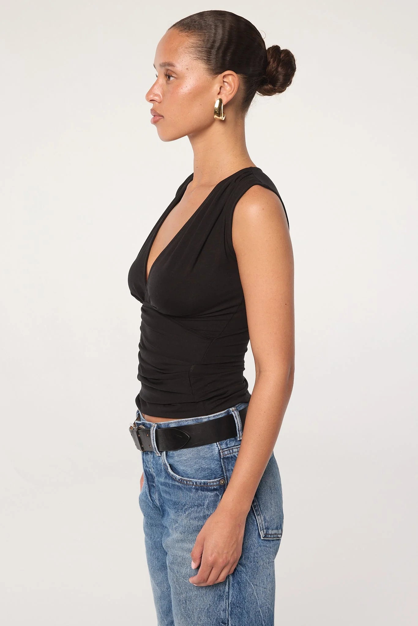 A woman in profile wears the Sunnery Top—a black v-neck made from soft Tencel jersey—paired with blue jeans, a black belt, and gold hoop earrings. Her hair is slicked back in a low bun, set against a plain white background.