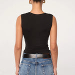 A person stands facing away, wearing the Sunnery Top—a black v-neck made from soft Tencel jersey—paired with blue jeans, a black belt, and gold hoop earrings. Their hair is styled in a neat bun against a plain, light-colored background.
