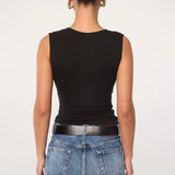 A person stands facing away, wearing the Sunnery Top—a black v-neck made from soft Tencel jersey—paired with blue jeans, a black belt, and gold hoop earrings. Their hair is styled in a neat bun against a plain, light-colored background.