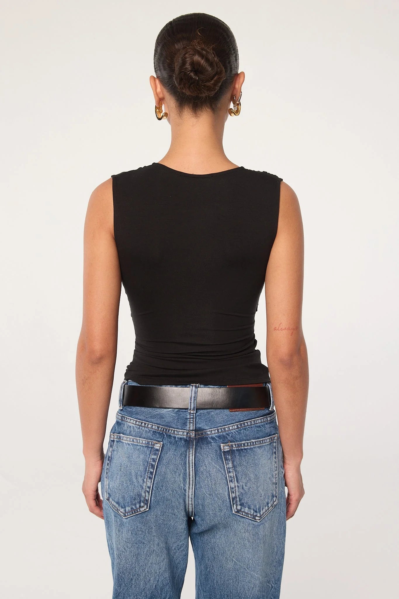 A person stands facing away, wearing the Sunnery Top—a black v-neck made from soft Tencel jersey—paired with blue jeans, a black belt, and gold hoop earrings. Their hair is styled in a neat bun against a plain, light-colored background.